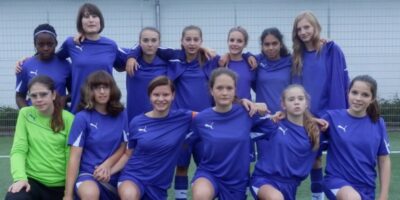 Eine Gruppe von Fußballspielerinnen posiert für ein Mannschaftsfoto. Alle tragen einheitliche, blaue Trikots und stehen vor einem Maschendrahtzaun. Einige Spielerinnen knien in der vorderen Reihe, während andere in der hinteren Reihe stehen und sich gegenseitig umarmen.