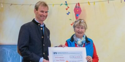 Eine Frau und ein Mann halten gemeinsam einen großen symbolischen Scheck über 2.100 Euro. Im Hintergrund sind bunte Dekorationen an einer gelben Wand zu sehen. Die Szene vermittelt eine freundliche und feierliche Atmosphäre.