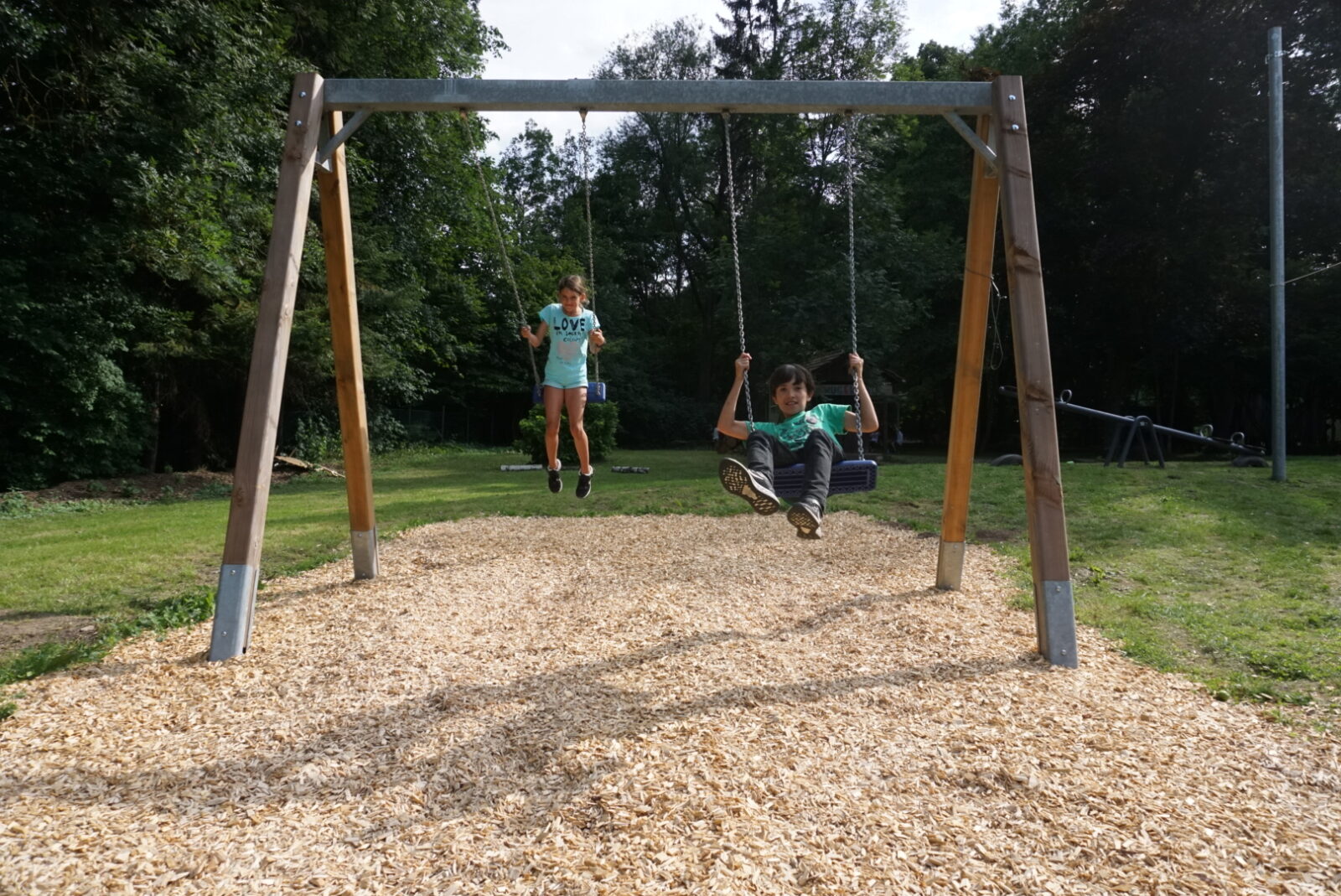 Ein Kind und eine Frau schaukeln auf einem Spielplatz umgeben von grünen Bäumen. Der Boden unter der Schaukel ist mit Holzschnitzeln bedeckt. Im Hintergrund sind weitere Spielgeräte und eine Wiese zu sehen.
