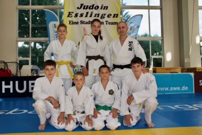 Eine Gruppe von Judokas posiert in ihren weißen Judo-Gis auf einer blauen Matte in einer Sporthalle. Im Hintergrund ist ein Banner mit der Aufschrift "Judo in Esslingen - Eine Stadt, ein Team" zu sehen. Die Sportler tragen Gürtel in verschiedenen Farben, die ihre jeweiligen Rangstufen im Judo anzeigen.