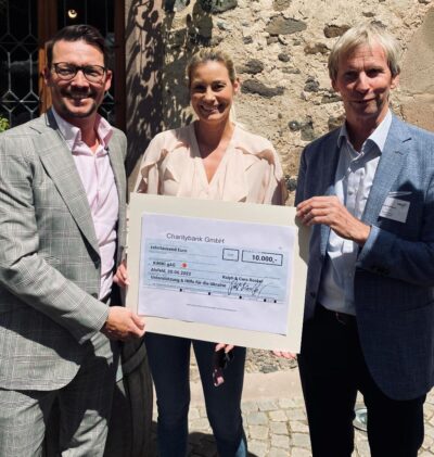 Drei Personen stehen nebeneinander und halten einen großen Scheck über 10.000 Euro von der Charitybank GmbH. Der Scheck ist für Unterstützung und Hilfe für die Ukraine datiert auf den 20.06.2022. Sie stehen vor einer Steinmauer und sind formell gekleidet.