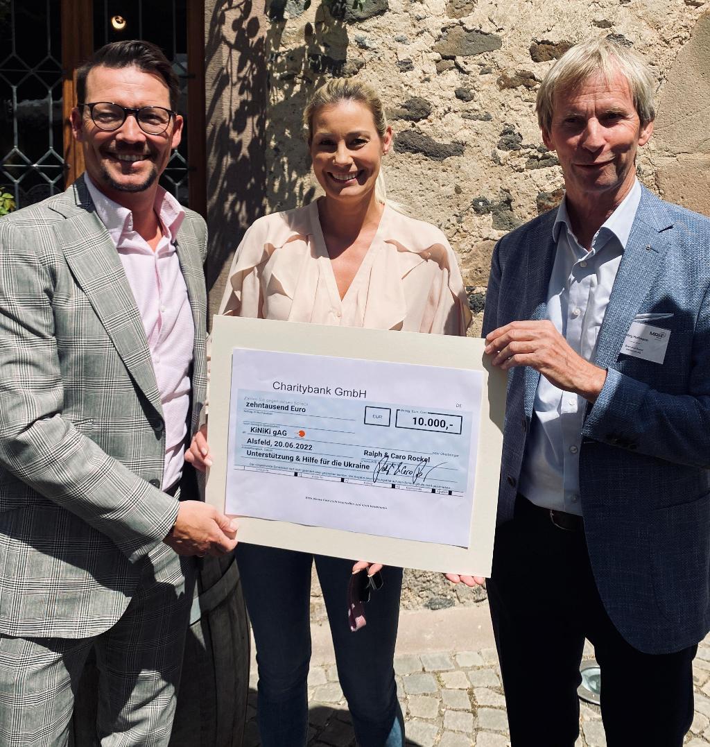 Drei Personen stehen nebeneinander und halten einen großen Scheck über 10.000 Euro von der Charitybank GmbH. Der Scheck ist für Unterstützung und Hilfe für die Ukraine datiert auf den 20.06.2022. Sie stehen vor einer Steinmauer und sind formell gekleidet.