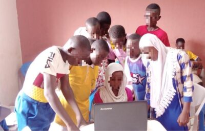 Eine Gruppe von Kindern versammelt sich um einen Laptop und schaut gespannt auf den Bildschirm. Die Kinder tragen bunte Kleidung und befinden sich in einem Raum mit einer rosafarbenen Wand. Der Laptop ist ein Lenovo-Gerät und scheint das Interesse der Gruppe zu wecken.
