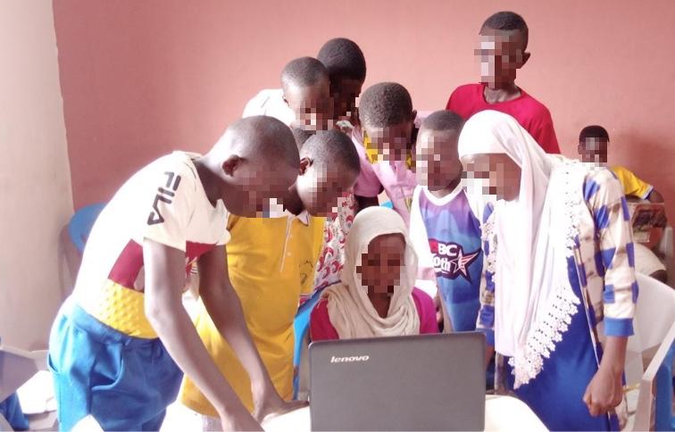 Eine Gruppe von Kindern versammelt sich um einen Laptop und schaut gespannt auf den Bildschirm. Die Kinder tragen bunte Kleidung und befinden sich in einem Raum mit einer rosafarbenen Wand. Der Laptop ist ein Lenovo-Gerät und scheint das Interesse der Gruppe zu wecken.