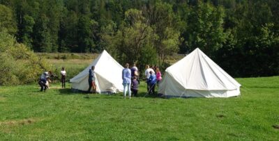 Eine Gruppe von Menschen baut zwei große weiße Zelte auf einer grünen Wiese auf. Im Hintergrund ist ein Wald zu sehen, der die Szene umrahmt. Die Personen arbeiten gemeinsam und scheinen sich in einer ländlichen Umgebung zu befinden.
