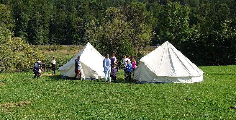 Eine Gruppe von Menschen baut zwei große weiße Zelte auf einer grünen Wiese auf. Im Hintergrund ist ein Wald zu sehen, der die Szene umrahmt. Die Personen arbeiten gemeinsam und scheinen sich in einer ländlichen Umgebung zu befinden.