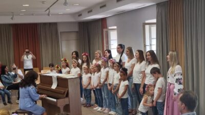 Eine Gruppe von Kindern und Erwachsenen steht nebeneinander in einem Raum und trägt weiße Oberteile und blaue Jeans. Einige der Kinder tragen bunte Blumenkränze auf ihren Köpfen. Eine Frau spielt Klavier, während die Gruppe aufmerksam zuhört. Im Hintergrund sind Zuschauer zu sehen, die das Geschehen beobachten und fotografieren.