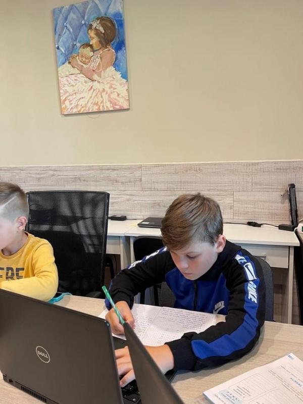 Zwei Kinder sitzen an einem Tisch und arbeiten konzentriert an ihren Aufgaben. Vor ihnen stehen Laptops und sie schreiben in Notizbücher. Im Hintergrund hängt ein farbenfrohes Gemälde an der Wand.