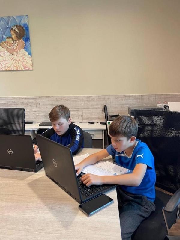 Zwei Kinder sitzen an einem Tisch und arbeiten konzentriert an ihren Laptops. Beide tragen sportliche Kleidung und haben Notizbücher neben sich liegen. Im Hintergrund ist eine Wand mit einem Gemälde zu sehen.