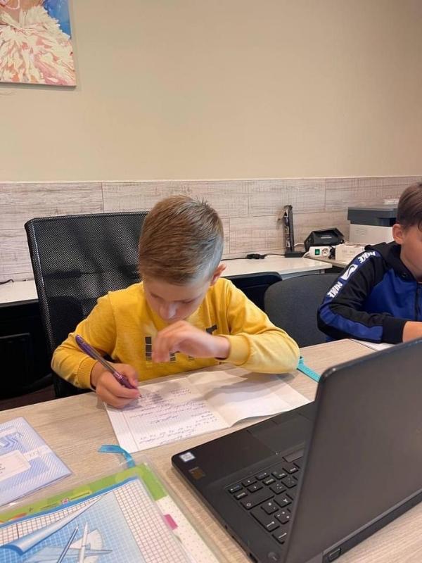 Ein Kind in einem gelben Pullover sitzt an einem Schreibtisch und schreibt auf ein Blatt Papier. Vor ihm steht ein Laptop, und auf dem Tisch liegen weitere Schulmaterialien. Im Hintergrund sind Büromaterialien und ein weiterer Schüler zu sehen.