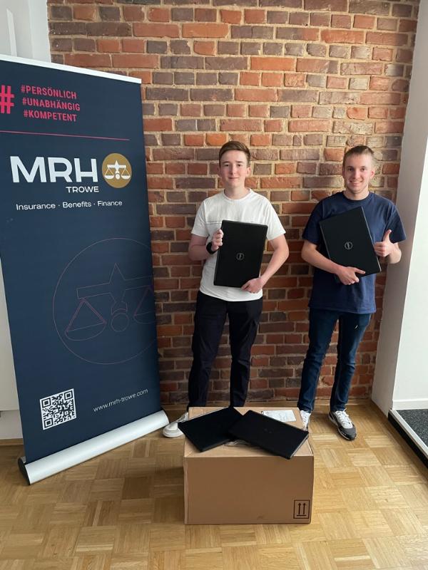 Zwei Personen stehen vor einer Backsteinwand und halten Ordner in den Händen. Neben ihnen ist ein Roll-Up-Banner von MRH Trowe zu sehen, das die Dienstleistungen des Unternehmens wie Versicherung, Benefits und Finanzen bewirbt. Vor den Personen steht ein großer Karton auf dem Boden.
