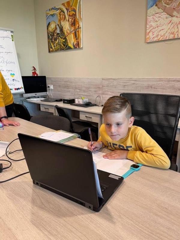Ein Kind sitzt an einem Tisch und arbeitet konzentriert mit einem Laptop und Notizbuch. Im Hintergrund sind ein Whiteboard und zwei Wandbilder zu sehen. Der Raum scheint ein Arbeits- oder Lernbereich zu sein.