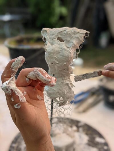 Zwei Hände formen eine Skulptur aus Draht und Ton. Die Finger sind mit Ton bedeckt, während die Skulptur langsam Gestalt annimmt. Im Hintergrund sind Werkzeuge und Materialien für die Bildhauerei zu sehen.