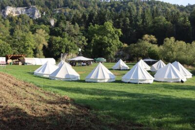 Eine Gruppe von weißen Zelten steht auf einer grünen Wiese, umgeben von üppigen Bäumen und einem bewaldeten Hügel im Hintergrund. Im Zentrum des Bildes ist ein größeres Pavillon-Zelt zu sehen, unter dem sich mehrere Menschen versammeln. Die Szene wirkt friedlich und einladend, ideal für ein Outdoor-Camping-Erlebnis.
