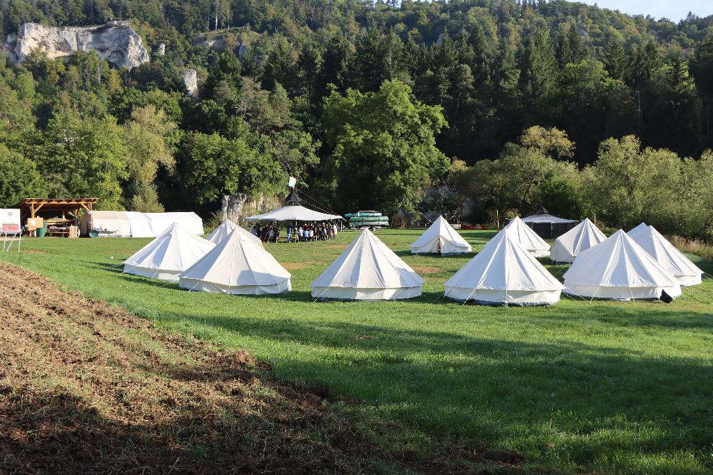Eine Gruppe von weißen Zelten steht auf einer grünen Wiese, umgeben von üppigen Bäumen und einem bewaldeten Hügel im Hintergrund. Im Zentrum des Bildes ist ein größeres Pavillon-Zelt zu sehen, unter dem sich mehrere Menschen versammeln. Die Szene wirkt friedlich und einladend, ideal für ein Outdoor-Camping-Erlebnis.