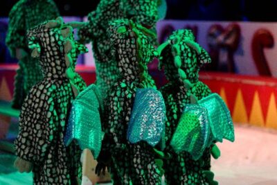 Kinder in Drachenkostümen stehen zusammen in einer bunten Umgebung, die an einen Zirkus erinnert. Die Kostüme sind grün und schwarz gemustert und haben schimmernde, blaue Flügel. Die Szene ist lebhaft beleuchtet und vermittelt eine fröhliche Atmosphäre.