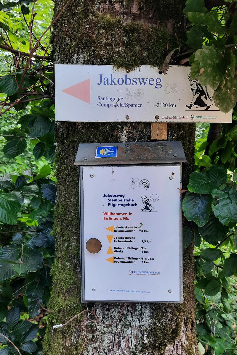 Ein Schild am Baum markiert den Jakobsweg mit einer Entfernung von 2120 km bis Santiago de Compostela in Spanien. Darunter befindet sich eine Stempelstelle für das Pilgertagebuch und Informationen zur Umgebung in Eislingen/Fils. Das Schild ist von grünen Blättern umgeben und bietet Orientierung für Wanderer und Pilger.