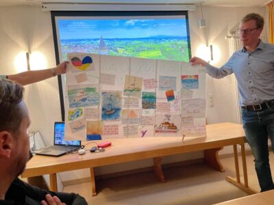Zwei Personen halten ein großes Poster mit verschiedenen Zeichnungen und Texten, das den Titel "Vom Segeln inspiriert" trägt. Im Hintergrund ist eine Präsentation mit einem Landschaftsbild auf einem Bildschirm zu sehen. Auf dem Tisch vor den Personen liegen ein Laptop und einige Schreibutensilien.