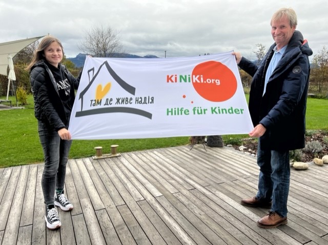 Zwei Personen halten ein Banner mit der Aufschrift "KiNiKi.org - Hilfe für Kinder" und einem Haus-Logo mit einem Herz, auf dem auch ukrainische Schriftzeichen zu sehen sind. Im Hintergrund befindet sich eine grüne Wiese und bewölkter Himmel. Das Bild vermittelt eine Botschaft der Unterstützung und Hilfe für Kinder.