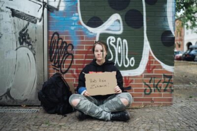 Eine Person sitzt auf dem Bürgersteig vor einer mit Graffiti bedeckten Wand und hält ein Schild mit der Aufschrift "Hab kein Zuhause!". Neben ihr liegt ein schwarzer Rucksack. Die Szene zeigt eine urbane Umgebung mit gepflastertem Boden und parkenden Autos im Hintergrund.