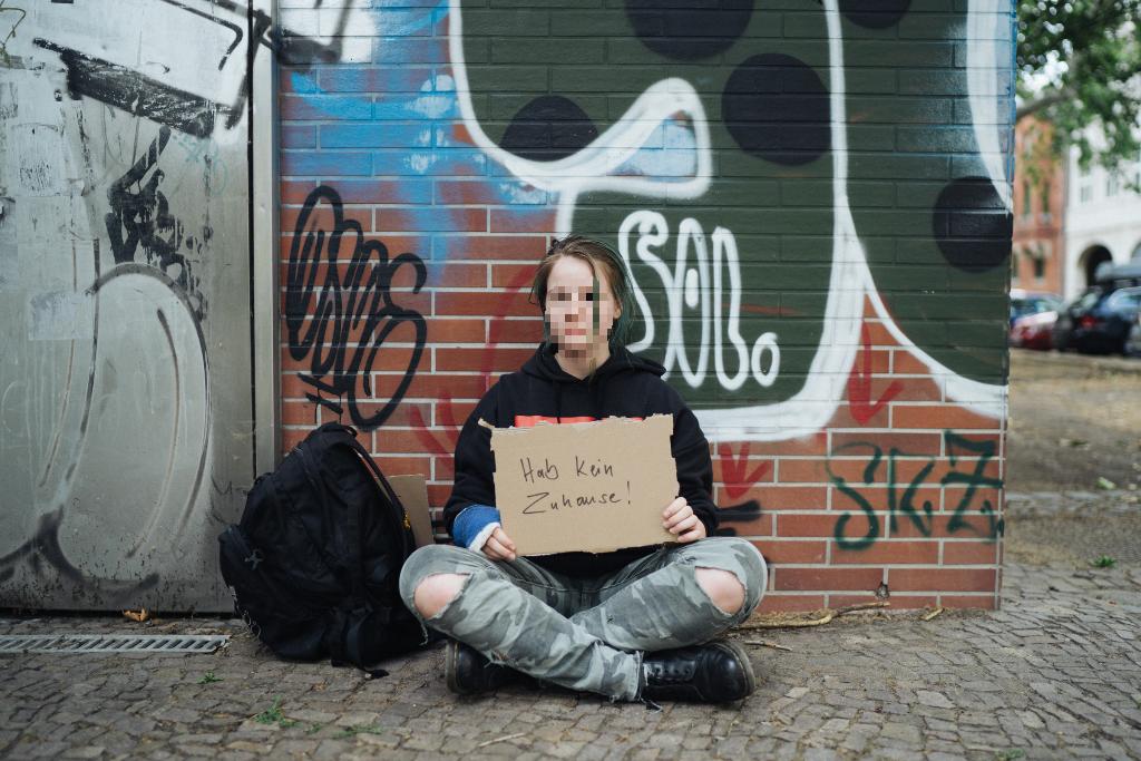 Eine Person sitzt auf dem Bürgersteig vor einer mit Graffiti bedeckten Wand und hält ein Schild mit der Aufschrift "Hab kein Zuhause!". Neben ihr liegt ein schwarzer Rucksack. Die Szene zeigt eine urbane Umgebung mit gepflastertem Boden und parkenden Autos im Hintergrund.