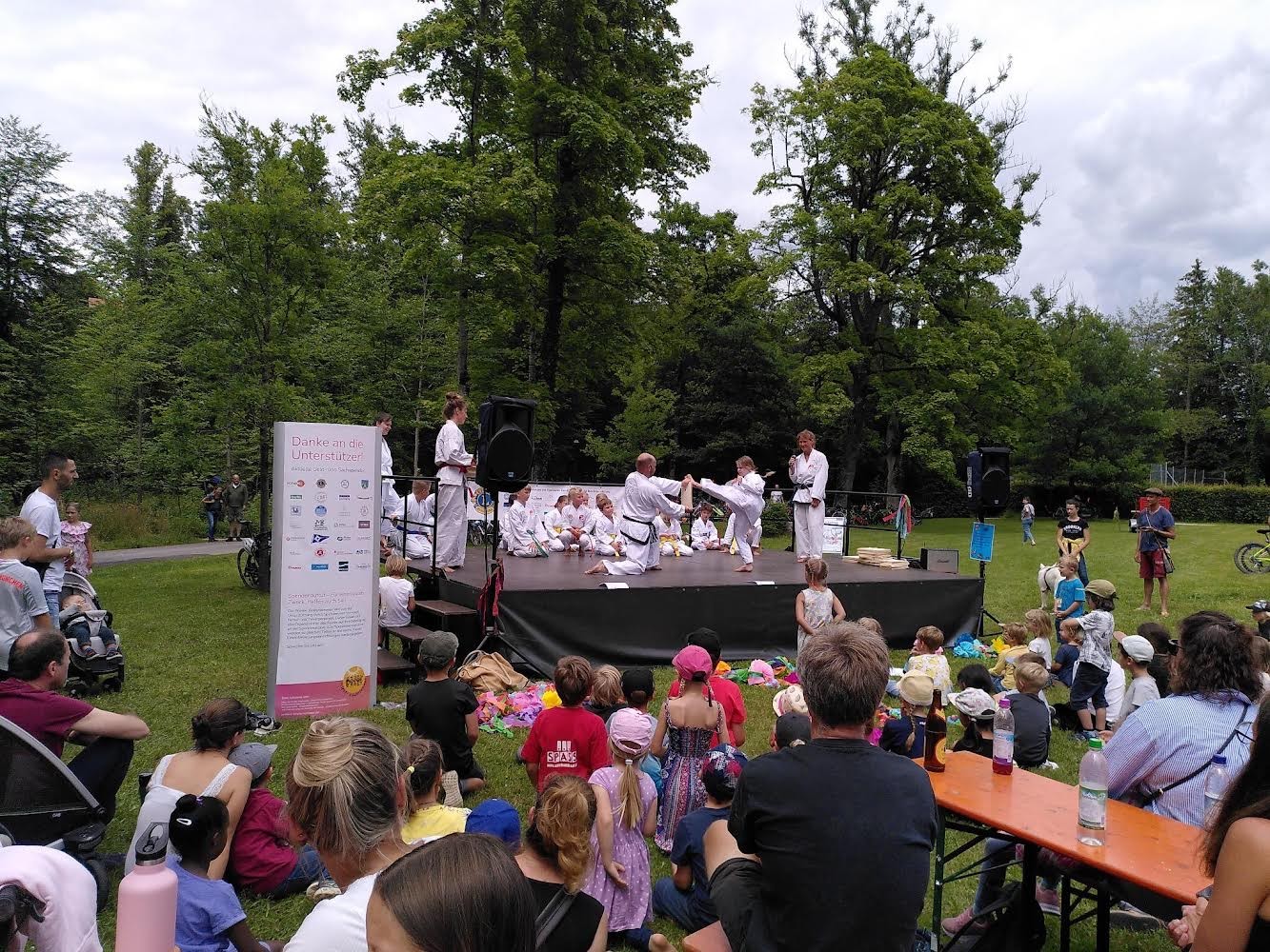 Eine Gruppe von Menschen versammelt sich im Freien, um eine Kampfkunstvorführung auf einer Bühne zu beobachten. Die Zuschauer, darunter viele Kinder, sitzen auf dem Gras und an Tischen, während die Darsteller in weißen Kampfanzügen auf der Bühne ihre Fähigkeiten demonstrieren. Im Hintergrund sind grüne Bäume und ein bewölkter Himmel zu sehen.