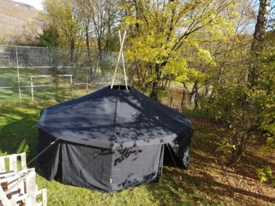 Ein großes schwarzes Zelt steht auf einer grünen Wiese, umgeben von Bäumen mit Herbstlaub. Im Hintergrund ist ein Maschendrahtzaun zu sehen, der ein Fußballfeld abgrenzt. Die Sonne scheint und wirft Schatten auf das Zelt und die Umgebung.