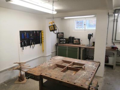 Eine gut ausgestattete Werkstatt mit einem großen Arbeitstisch im Vordergrund. An der Wand hängen verschiedene Werkzeuge, darunter Schraubenzieher und Zangen, ordentlich aufgereiht. Im Hintergrund sind ein Bohrgerät und mehrere Werkzeugkisten auf einer Arbeitsfläche zu sehen, die unter einem Fenster platziert ist.