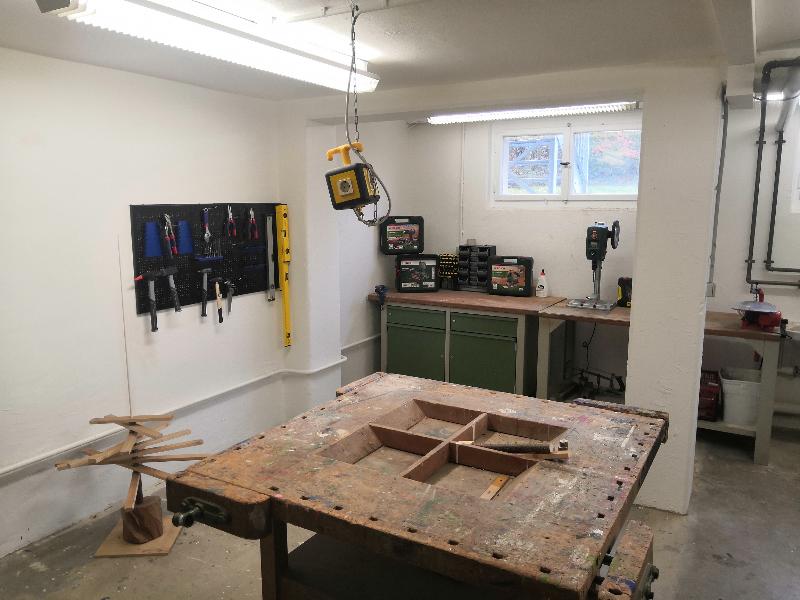 Eine gut ausgestattete Werkstatt mit einem großen Arbeitstisch im Vordergrund. An der Wand hängen verschiedene Werkzeuge, darunter Schraubenzieher und Zangen, ordentlich aufgereiht. Im Hintergrund sind ein Bohrgerät und mehrere Werkzeugkisten auf einer Arbeitsfläche zu sehen, die unter einem Fenster platziert ist.