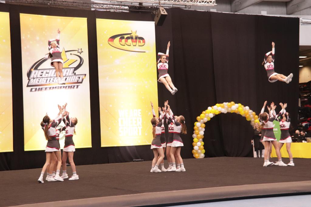 Ein Cheerleading-Team führt eine beeindruckende Hebefigur während eines Wettbewerbs auf. Die Cheerleader sind in der Luft und werden von ihren Teamkollegen unterstützt. Im Hintergrund sind gelbe Banner und ein Ballonbogen zu sehen, die die Veranstaltung dekorieren.
