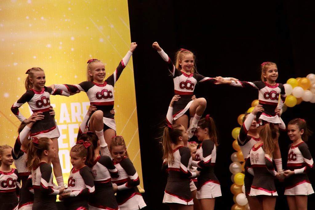 Eine Gruppe von Cheerleadern führt eine akrobatische Pyramide vor, wobei einige Mitglieder auf den Schultern ihrer Teamkollegen stehen. Die Cheerleader tragen einheitliche Outfits in Schwarz, Weiß und Rot mit dem Logo "CC" auf der Vorderseite. Im Hintergrund ist ein gelbes Banner mit der Aufschrift "ARE YOU READY" zu sehen.