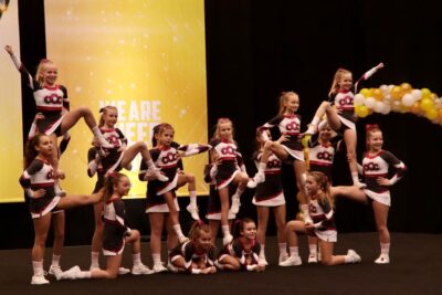 Eine Gruppe von Cheerleadern führt eine beeindruckende Pyramidenformation auf einer Bühne auf. Sie tragen einheitliche rot-weiß-schwarze Kostüme und zeigen verschiedene akrobatische Posen. Im Hintergrund sind gelbe Banner und ein Bogen aus Luftballons zu sehen.