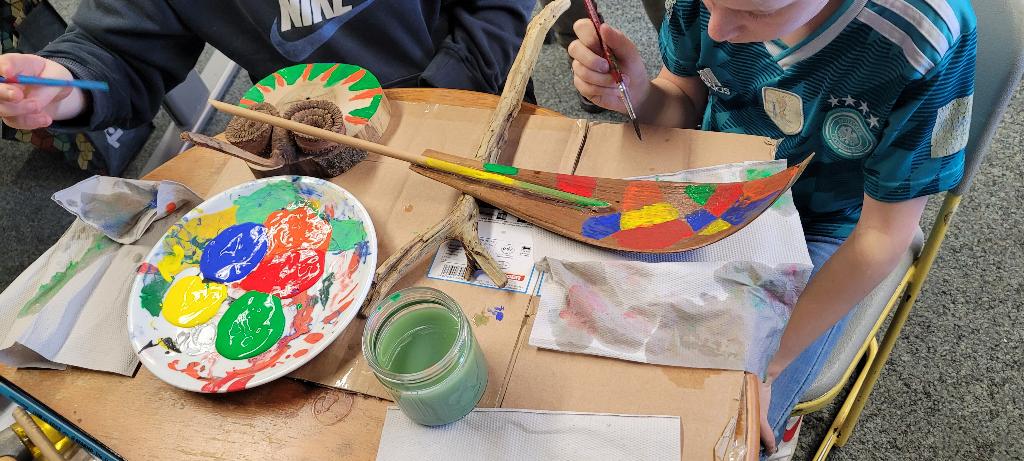 Zwei Personen arbeiten an einem Bastelprojekt und bemalen ein hölzernes Modellboot mit bunten Farben. Auf dem Tisch liegen verschiedene Bastelmaterialien, darunter eine Farbpalette mit lebhaften Farben und ein Glas mit grünem Wasser. Die Szene zeigt kreative Handarbeit und gemeinschaftliches Basteln.