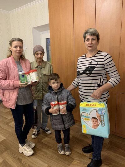 Eine Gruppe von Menschen steht in einem Raum und hält verschiedene Babyprodukte, darunter Milchpulver und eine große Packung Windeln. Sie sind warm gekleidet und wirken wie eine Familie, die sich um die Versorgung eines Kindes kümmert. Die Szene vermittelt einen Eindruck von Fürsorge und Gemeinschaft.