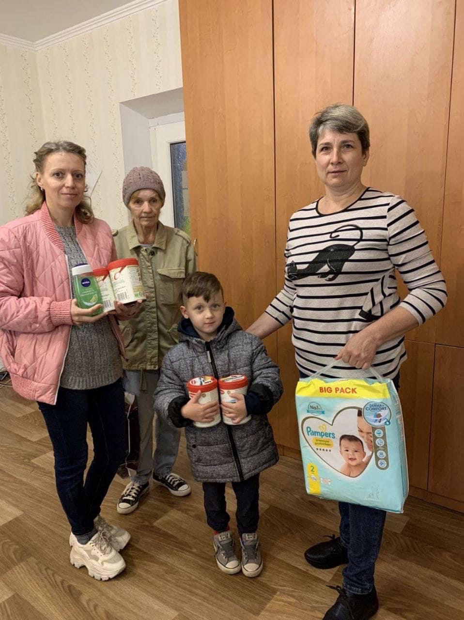 Eine Gruppe von Menschen steht in einem Raum und hält verschiedene Babyprodukte, darunter Milchpulver und eine große Packung Windeln. Sie sind warm gekleidet und wirken wie eine Familie, die sich um die Versorgung eines Kindes kümmert. Die Szene vermittelt einen Eindruck von Fürsorge und Gemeinschaft.
