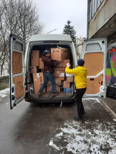 Zwei Männer entladen einen Lieferwagen, der mit zahlreichen Kartons gefüllt ist. Einer der Männer trägt eine gelbe Jacke mit der Aufschrift "FC SKELLE" und steht im Schnee, während der andere Mann im Inneren des Lieferwagens steht und einen Karton heraushebt. Im Hintergrund sind kahle Bäume und Gebäude zu sehen, die auf eine winterliche Umgebung hinweisen.