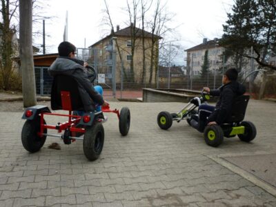 Zwei Kinder fahren auf einem gepflasterten Weg in einem Park auf Go-Karts. Im Hintergrund sind Bäume, ein Zaun und Gebäude zu sehen. Beide Kinder tragen warme Kleidung und scheinen Spaß zu haben.