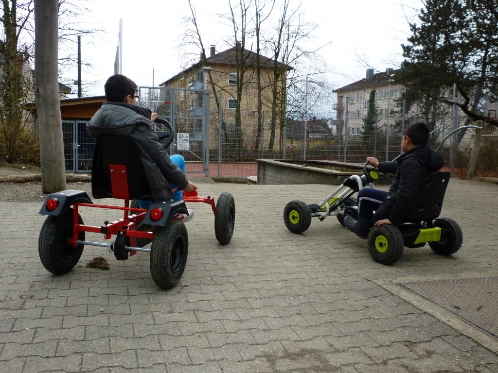 Zwei Kinder fahren auf einem gepflasterten Weg in einem Park auf Go-Karts. Im Hintergrund sind Bäume, ein Zaun und Gebäude zu sehen. Beide Kinder tragen warme Kleidung und scheinen Spaß zu haben.