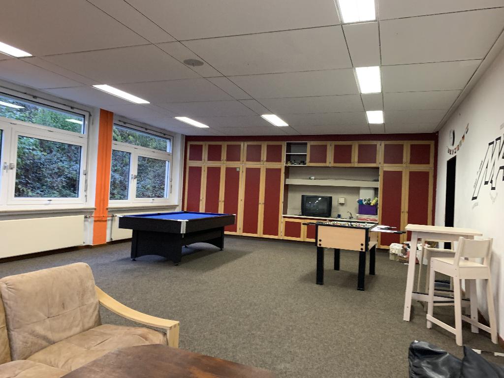 Ein Freizeitraum mit einem Billardtisch und einem Tischfußballspiel in der Mitte. An der Wand befinden sich rote und gelbe Schränke, und ein Fernseher steht auf einem Regal. Große Fenster lassen viel Tageslicht herein und bieten einen Blick ins Grüne.