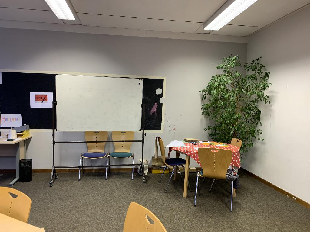 Ein Klassenzimmer mit einer großen weißen Tafel und mehreren Stühlen. Rechts im Bild steht ein Tisch mit einer rot-weiß karierten Tischdecke und einigen Büchern darauf. Eine grüne Zimmerpflanze befindet sich in der Ecke des Raumes.