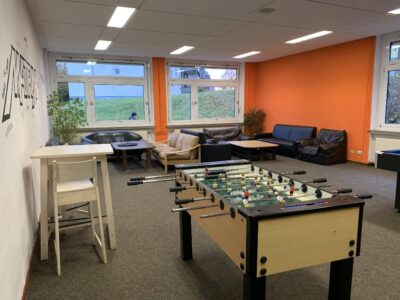 Ein Gemeinschaftsraum mit einem Kickertisch im Vordergrund. Im Hintergrund befinden sich Sofas und Sitzgelegenheiten vor einer orangefarbenen Wand und großen Fenstern, die einen Blick ins Freie bieten. Eine gemütliche Atmosphäre lädt zum Entspannen und Spielen ein.