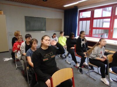 Eine Gruppe von Schülerinnen und Schülern sitzt in einem Klassenzimmer auf Stühlen, die in zwei Reihen angeordnet sind. Im Hintergrund sind eine grüne Tafel und ein Fenster mit roten Rahmen zu sehen. Einige der Schülerinnen und Schüler tragen sportliche Kleidung und scheinen miteinander zu interagieren.