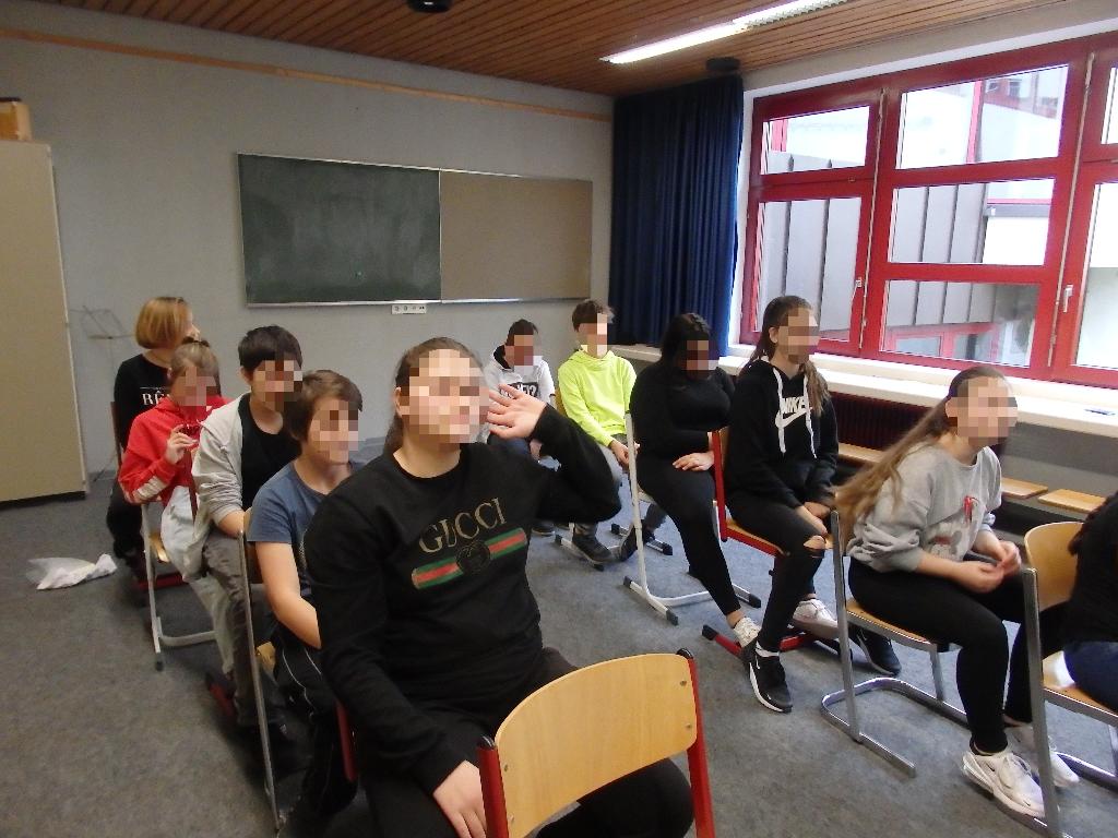 Eine Gruppe von Schülerinnen und Schülern sitzt in einem Klassenzimmer auf Stühlen, die in zwei Reihen angeordnet sind. Im Hintergrund sind eine grüne Tafel und ein Fenster mit roten Rahmen zu sehen. Einige der Schülerinnen und Schüler tragen sportliche Kleidung und scheinen miteinander zu interagieren.