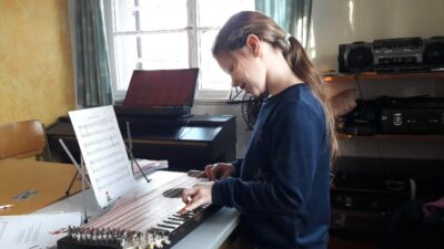 Eine Person sitzt an einem Klavier und spielt darauf. Der Raum ist hell und gemütlich eingerichtet, mit einem Fenster im Hintergrund.