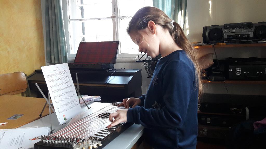 Eine Person sitzt an einem Klavier und spielt darauf. Der Raum ist hell und gemütlich eingerichtet, mit einem Fenster im Hintergrund.