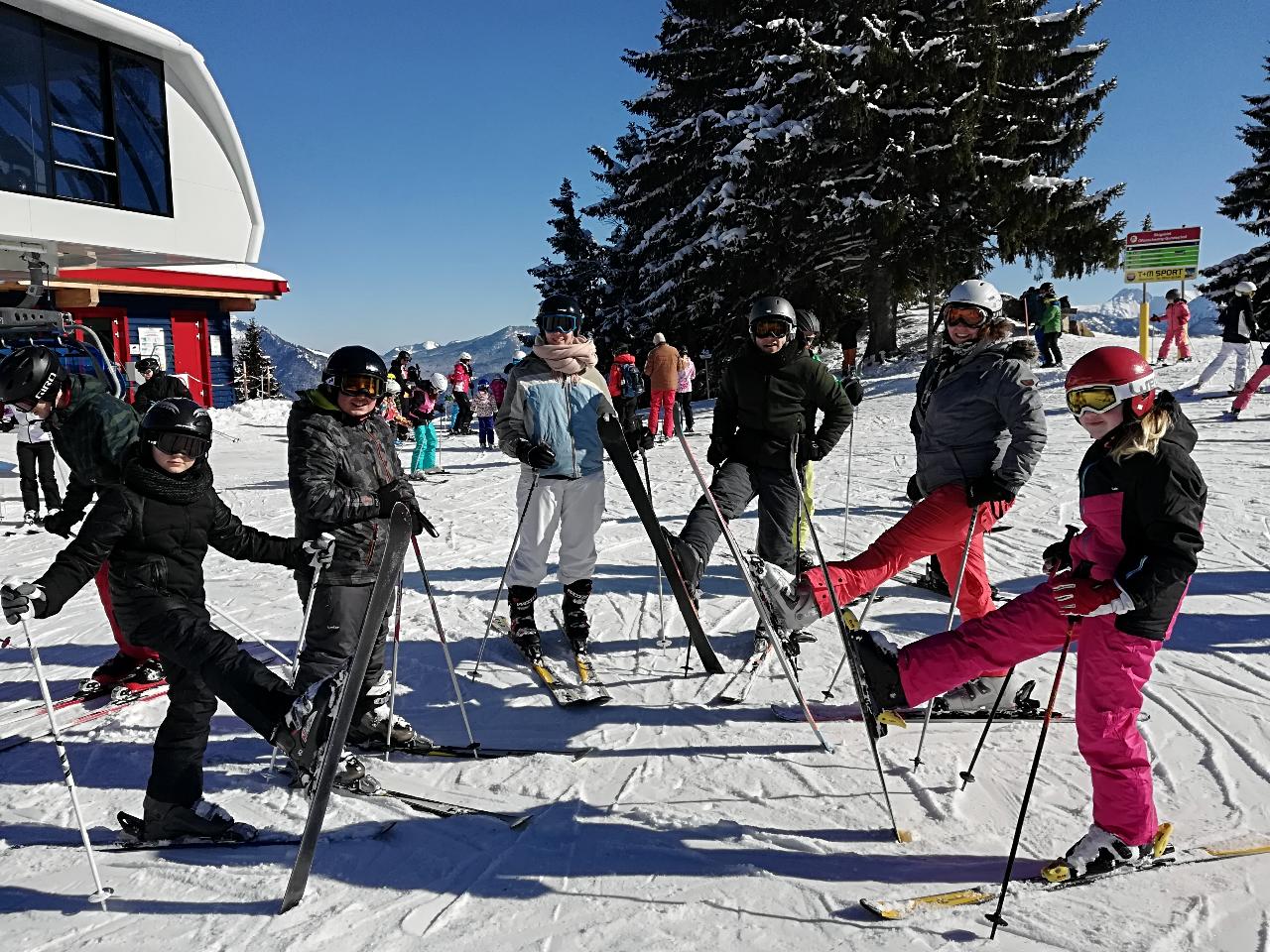 Eine Gruppe von Menschen steht auf einer Skipiste und bereitet sich auf das Skifahren vor. Sie tragen warme Kleidung und Skiausrüstung, während im Hintergrund verschneite Bäume und Gebäude zu sehen sind.
