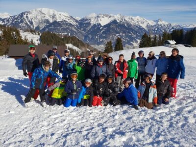 Eine Gruppe von Menschen steht in winterlicher Kleidung auf einem schneebedeckten Berg. Im Hintergrund sind weitere schneebedeckte Berge und ein klarer Himmel zu sehen.