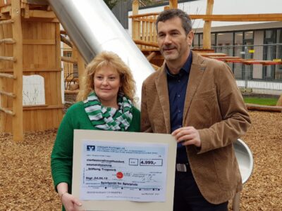 Zwei Personen halten gemeinsam einen Scheck. Sie stehen vor einem Spielplatz, der im Hintergrund zu sehen ist.