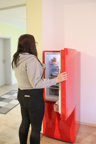 Eine Person steht vor einem roten Automaten und scheint ihn zu bedienen. Der Automat steht in einem hellen Raum mit kariertem Boden.