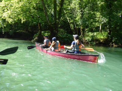 Drei Personen paddeln in einem roten Kanu auf einem ruhigen Fluss, umgeben von üppigem grünen Wald. Sie tragen Schwimmwesten und genießen offensichtlich die friedliche Natur. Das Sonnenlicht fällt durch die Bäume und erzeugt ein wunderschönes Spiel von Licht und Schatten auf dem Wasser.